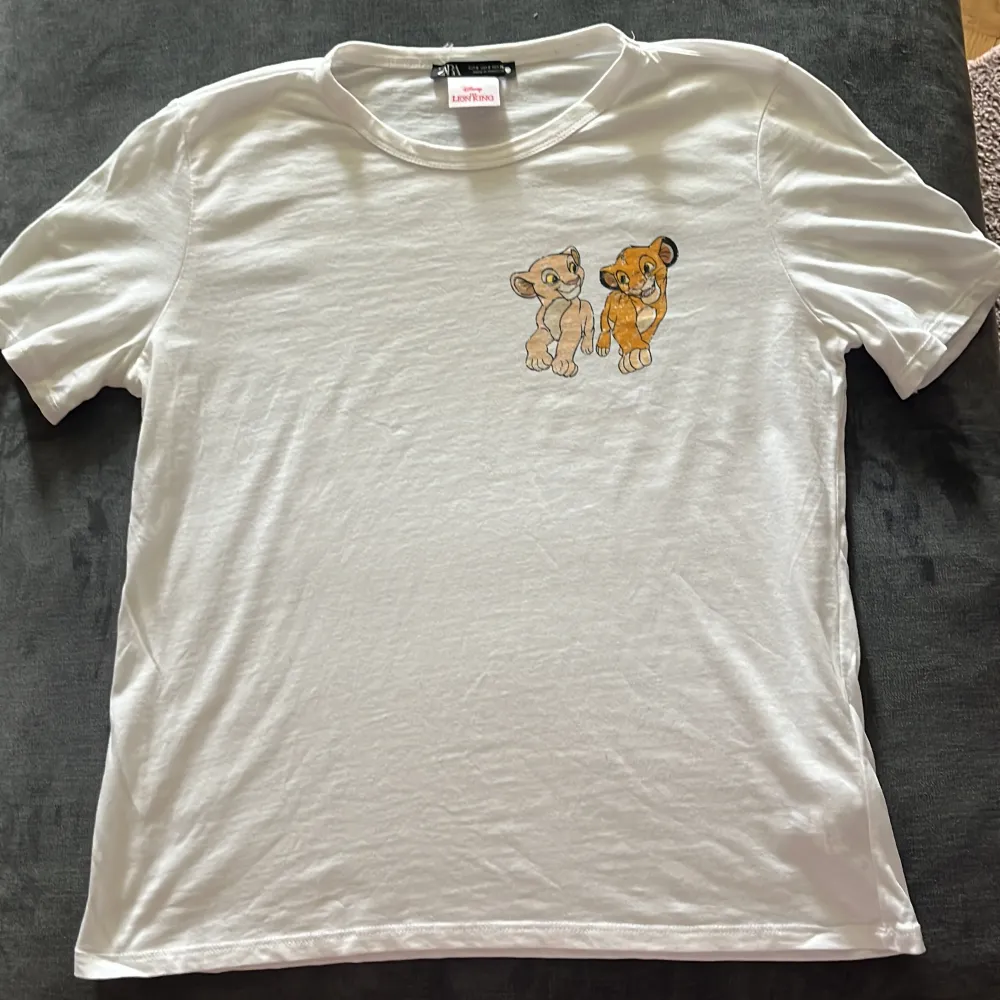 Säljer en superfin vit t-shirt från Zara med ett gulligt Lejonkungen-tryck på bröstet. Perfekt för alla Disney-fans! T-shirten är i bomull och har en normal passform med korta ärmar. Perfekt för en avslappnad stil.. T-paidat.