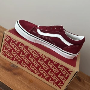 Vinröda sneakers från Vans - Snygga vinröda sneakers från Vans med klassisk vit sula och vit sidostrimma. Skorna har en låg profil och är tillverkade i mocka med snörning framtill. Perfekta för en avslappnad stil.