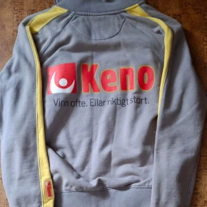 Grå och gul fleecejacka med Keno-tryck - Säljer en grå fleecejacka med gula detaljer och Keno-tryck. Jackan har dragkedja och långa ärmar. Perfekt för kyliga dagar!