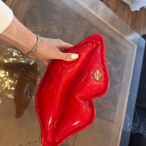Röd läppformad väska - Röd  lackväska i form av läppar med en glansig yta. Väskan har en dragkedja på baksidan för enkel öppning och stängning. Perfekt för att ge en färgklick till din outfit.