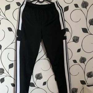 Svarta träningsbyxor från Adidas - Snygga svarta träningsbyxor från Adidas med vita ränder längs sidorna. Byxorna har en dragkedja vid bensluten och elastisk midja. Perfekta för träning eller en avslappnad stil. Är i barnstorlek men passar som XS.