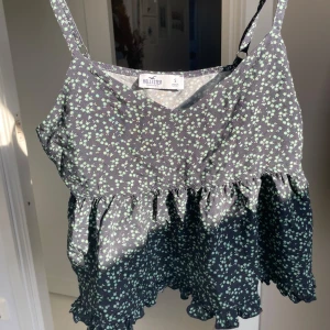 Blommig linnetopp från Hollister - Säljer en söt blommig linnetopp från Hollister, aldrig använd. 💞Toppen är svart med små gröna blommor och har justerbara axelband. Den har en volangdetalj vid nederkanten som ger en fin touch. Perfekt för varma dagar! Kan diskutera priset!🩷🩷🩷