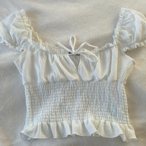 Vit topp från Gina Tricot - Säljer en söt vit topp från Gina Tricot med volanger och knytning framtill. Toppen är croppad och har korta ärmar med en smockad design som ger en fin passform. Perfekt för varma dagar!