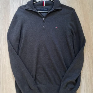 Grå tröja med dragkedja från Tommy Hilfiger - Säljer en stilren grå tröja från Tommy Hilfiger med dragkedja vid halsen. Tröjan har en liten logotyp på bröstet och är perfekt för en avslappnad look. Den är långärmad och tillverkad i ett mjukt material.