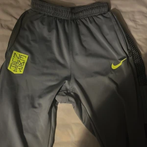 Gråa Nike underställ  - Snygga grå träningsbyxor från Nike med Dri-FIT-teknologi. Byxorna har en elastisk midja och dragkedjor vid fickorna. Dekorerade med en gul Nike-logga och Neymar-tryck på sidan. Perfekta för sport och aktivitet.