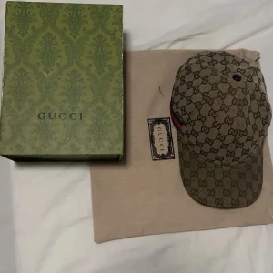 Beige keps från Gucci - Säljer en stilren beige keps från Gucci med det ikoniska GG-mönstret. Kepsen har en klassisk passform och är perfekt för att ge en lyxig touch till din outfit. Den kommer med originalförpackning och etikett. Storlek L (57 cm)