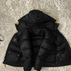 Svart dunjacka från Fjällräven - Säljer en svart dunjacka från Fjällräven Expedition Series. Jackan har en mysig huva och praktiska fickor med knappar. Den är perfekt för kalla dagar med sin varma vaddering och stilrena design. Gula detaljer ger en snygg kontrast. Perfekt för vintern! ❄️ OBS priset går att diskutera och kvitto finns