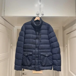 Mörkblå dunjacka från Herno - Säljer en stilren mörkblå dunjacka från Herno. Jackan har en hög krage och praktiska fickor med knappar framtill. Den är tillverkad med innovativ dunteknik för lätt och mjuk känsla. Perfekt för kyligare dagar. Finns små små knappt synliga hål som jag kan skicka bild på om man är intresserad 