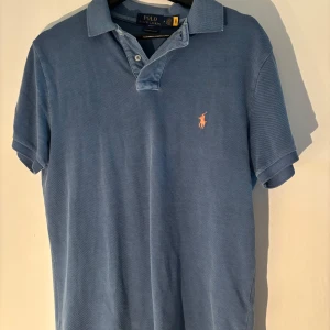 Blå pikétröja från Ralph Lauren - Säljer en stilren blå pikétröja från Ralph Lauren i storlek M. Tröjan har en klassisk krage och korta ärmar med en broderad logotyp i orange på bröstet. Perfekt för en avslappnad men ändå snygg look.
