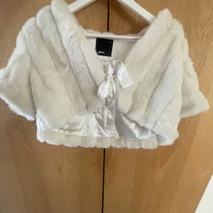 Vit pälsbolero från Gina - Säljer en elegant vit pälsbolero från Gina med korta ärmar och en söt knytning framtill. Perfekt för att lägga till en touch av glamour till din outfit. Jackan har en mjuk och lyxig känsla.Priset går att diskutera!💗💗