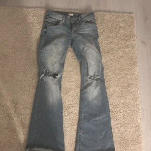 Blå bootcut jeans med slitningar - Snygga blå bootcut jeans med slitningar på knäna. Perfekta för en avslappnad stil. De har en klassisk femficksdesign och en knappgylf. Jeansen har en ljus tvätt som ger en vintagekänsla.
