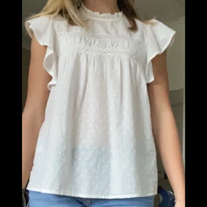 Vit blus med spetsdetaljer - Säljer denna söta vita blus med spetsdetaljer och volanger på ärmarna. Blusen har ett diskret prickigt mönster och en knapp i nacken.🩷🩷