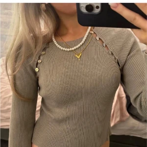 Beige ribbad tröja med knappar - Snygg beige ribbad tröja med unika knappar längs axeln. Tröjan har en tight passform och långa ärmar, perfekt för en stilren look. Passar bra till både vardag och speciella tillfällen.