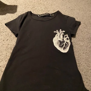 Svart t-shirt med hjärtmotiv - Snygg svart t-shirt från Lager 157 med ett unikt hjärtmotiv tryckt på bröstet. Perfekt för en cool och avslappnad stil. Kortärmad och tillverkad i mjukt material för extra komfort.