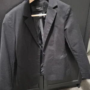 Svart kavaj från Vero Moda - Snygg svart kavaj från Vero Moda i storlek M. Perfekt för en stilren look med klassisk design och två knappar framtill. Passar utmärkt till både vardag och festliga tillfällen.