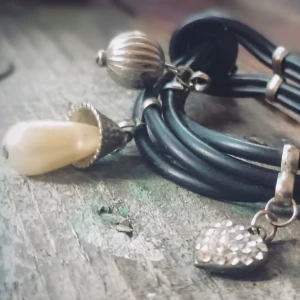 Svart läderarmband med pärldetaljer - Elegant svart läderarmband med flera remmar och dekorativa detaljer. Armbandet har en vit pärla och ett hjärta med glittriga stenar som hänger som berlocker. Perfekt för att ge en stilfull touch till din outfit.