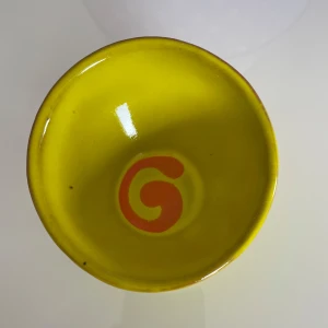 Gul keramikskål med orange mönster - Charmig gul keramikskål med ett unikt orange mönster i botten. Skålen är handgjord i Grekland och har en rund form med en blank yta. Perfekt för att ge en färgglad touch till ditt kök eller vardagsrum..namn glentzanis . Storlek på höjd är 7cm och rund 9,05cm 
