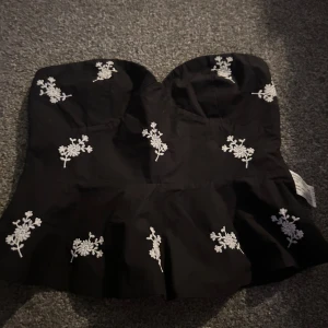 Svart axelbandslös topp med broderi - Snygg svart axelbandslös topp från Zara med vita blombroderier. Toppen har en volangdetalj vid nederkanten och en smockad rygg för bättre passform. Perfekt för en stilren look.