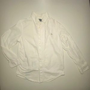 Vit skjorta från Ralph Lauren - Klassisk vit skjorta från Ralph Lauren med broderad logotyp på bröstet. Skjortan har långa ärmar och knappar framtill, det sitter slim och passar perfekt för en stilren look. Jag på bilden är runt 174 och den är för liten för mig. Skjortan passar dig som är mellan 160-170. Priset kan diskuteras!