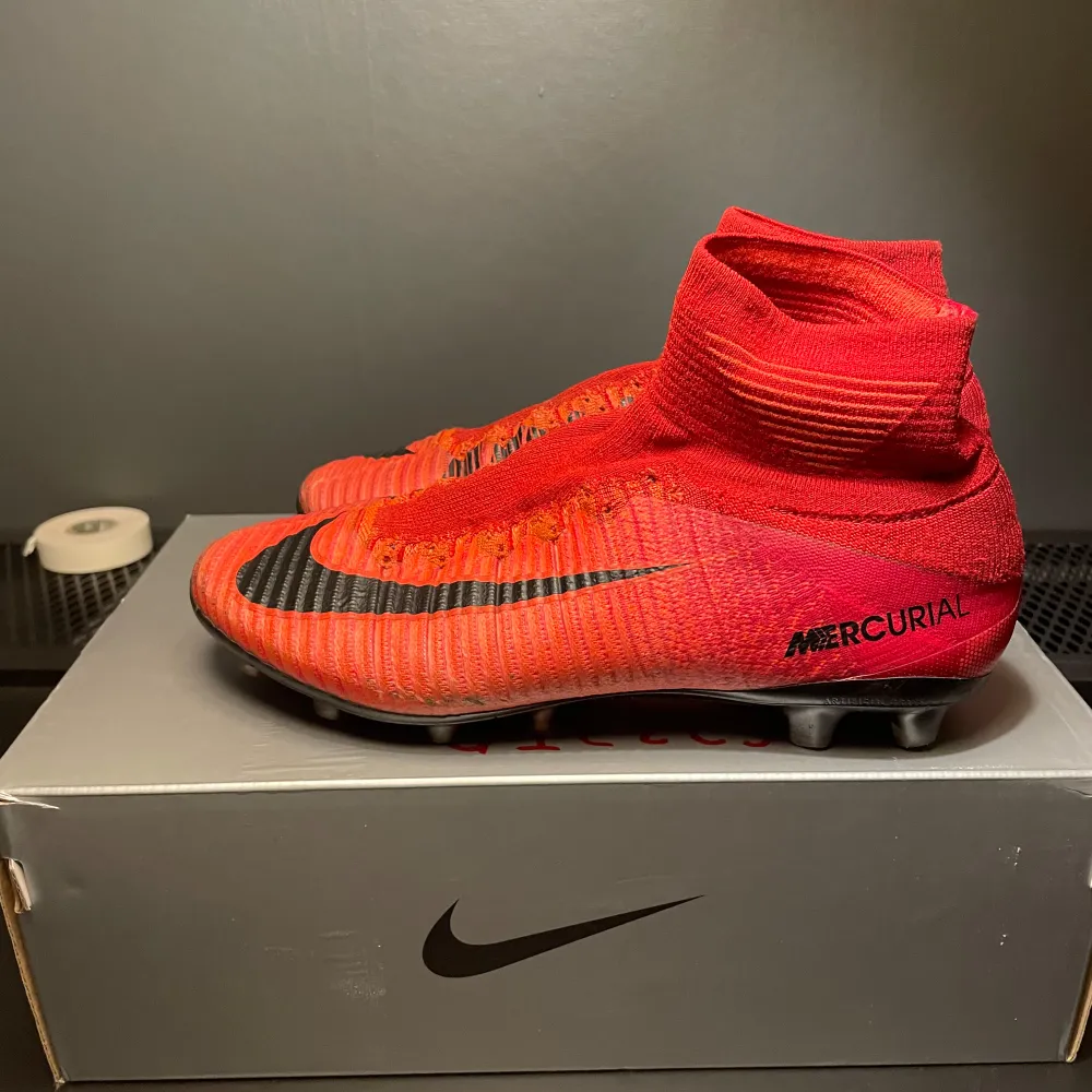 Säljer ett par röda Nike Mercurial fotbollsskor med hög ankelkrage och svart swoosh-logga. Skorna har en strömlinjeformad design och är perfekta för snabba rörelser på planen. De är designade för att ge bra grepp och kontroll.. Kengät.