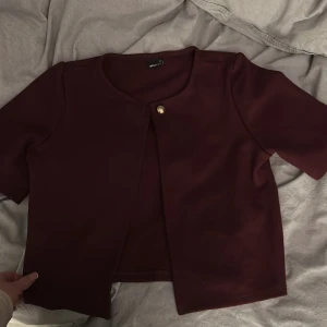 Vinröd kofta  - Säljer en stilren vinröd jacka från Gina Tricot med korta ärmar och en elegant knappdetalj framtill. Perfekt för att lägga till en touch av färg till din outfit. Passar bra till både vardag och speciella tillfällen.
