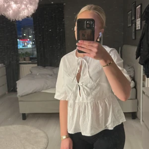 Vit blus från Divided - Säljer en söt vit blus från Divided med knytning framtill och korta ärmar. Perfekt för en stilren look. Blusen har en luftig passform och är tillverkad i ett lätt material.