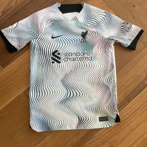 Vit och svart fotbollströja- Liverpool bortaställ 2022-2023 - Säljer en cool vit och svart fotbollströja från Nike med ett unikt vågmönster. Tröjan har korta ärmar och tryck med 'Darwin 27' på baksidan. Premier League-logga på ärmen. Perfekt för fotbollsfans!