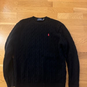 Svart stickad tröja från Polo Ralph Lauren - Säljer en klassisk svart stickad tröja från Polo Ralph Lauren med det ikoniska röda logotypbroderiet på bröstet. Tröjan har en rund halsringning och långa ärmar, perfekt för kyligare dagar. Litet hål vid slutet av ärmen som lätt går stt sy ihop
