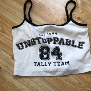 Vit och svart linne med tryck - Säljer en cool vit linne från Tally Weijl med svart text 'UNSTOPPABLE 84 TALLY TEAM' på framsidan. Den har svarta kanter och smala axelband. Perfekt för en avslappnad stil.