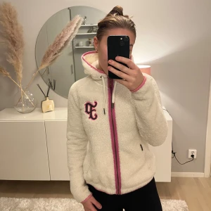 Vit fleecejacka från Peak Performance - Mysig vit fleecejacka från Peak Performance med rosa detaljer och dragkedja. Jackan har en broderad logga på bröstet och en praktisk huva. Perfekt för kyliga dagar när du vill hålla dig varm och bekväm eller på en skidresa.