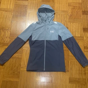 Grå vindjacka från Under Armour - Säljer en stilren grå vindjacka från Under Armour. Jackan har en justerbar huva och dragkedja framtill. Perfekt för blåsiga dagar med sin lätta och funktionella design. Logotypen är tryckt på bröstet. Vill få bort den så snabbt som möjligt!