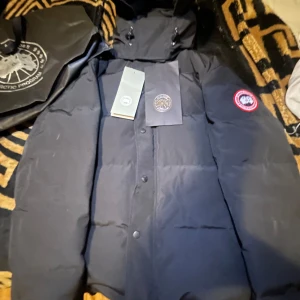 Svart dunjacka från Canada Goose - Säljer en svart dunjacka från Canada Goose med huva och knappar framtill. Jackan har en klassisk design med en rund logotyp på ärmen. Perfekt för kalla dagar.