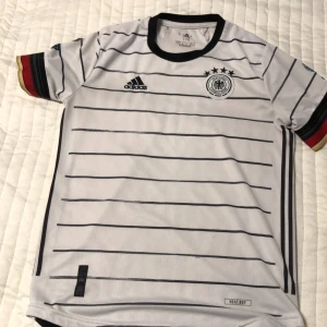 Vit fotbollströja från Adidas - Säljer en vit fotbollströja från Adidas med svarta ränder och Tysklands landslagsemblem. Tröjan har korta ärmar med röd och gul detalj vid ärmsluten. Perfekt för fotbollsfans! Nypris 1200