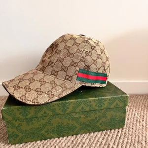 Beige keps från Gucci - Snygg beige keps från Gucci med det klassiska GG-mönstret. Kepsen har en grön och röd randdetalj på sidan. Perfekt för att ge din outfit en lyxig touch.