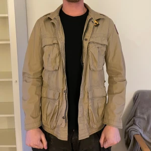 Parajumpers field jacket XL - Passar L och XL. Säljer en beige jacka från Parajumpers med flera praktiska fickor och en dragkedja framtill. Jackan har en broderad logga på ärmen och en klassisk krage. Perfekt för vår och höst!