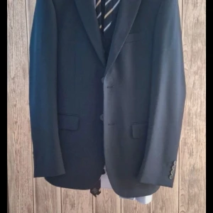 Svart kostym från Dressmann - Snygg svart kostym från Dressmann i slim fit. Kavajen har klassiska slag och knappar framtill. Västen matchar perfekt med kavajen och har knappar. Perfekt för formella tillfällen.