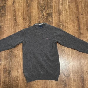 Grå ulltröja från GANT - Säljer en stilren grå ulltröja från GANT med rund halsringning och broderad logotyp på bröstet. Perfekt för kyligare dagar och passar till både jeans och chinos.