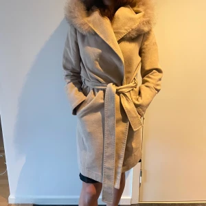 Beige kappa med pälskrage - Elegant beige kappa med en lyxig pälskrage och huva. Jackan har ett knytbälte i midjan för en smickrande passform och långa ärmar. Perfekt för kyliga dagar när du vill hålla stilen.