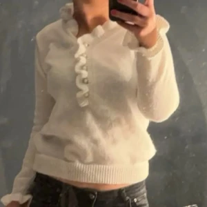 Vit stickad tröja med volangdetaljer - Säljer en söt vit stickad tröja med volangdetaljer längs knäppningen. Tröjan har långa ärmar och en mysig passform, perfekt för kyligare dagar.