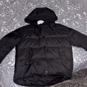 Svart pufferjacka med huva - Säljer en svart pufferjacka med huva från vero moda. Jackan har långa ärmar och stängs med knappar. Perfekt för kyligare dagar.