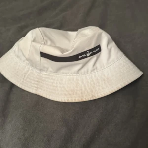 Vit hatt från Sail Racing - Snygg vit hatt från Sail Racing med svart logotypdetalj. Perfekt för soliga dagar och en stilren look. Hatten har en klassisk design med bred kant.