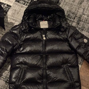 Svart dunjacka från Moncler - Säljer en snygg svart dunjacka från Moncler med huva och dragkedja. Jackan har en glansig finish och Moncler-logga på ärmen. Perfekt för kyliga dagar.