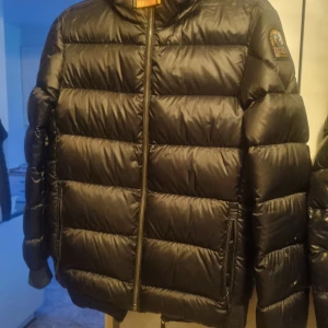 Svart dunjacka från Parajumpers st 164-170 - Snygg svart dunjacka från Parajumpers med dragkedja och huva. Jackan har en quiltad design och en patch på ärmen. Perfekt för kyliga dagar st 164-170 passar också till St S 