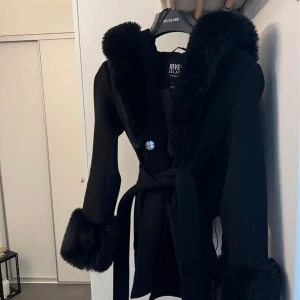 Svart kappa med fuskpäls från River Island - Elegant svart kappa från River Island med fuskpälsdetaljer på krage, ärmslut och huva. Kappan har ett knytbälte i midjan och en knapp framtill. Perfekt för kyliga dagar när du vill hålla stilen. St 34