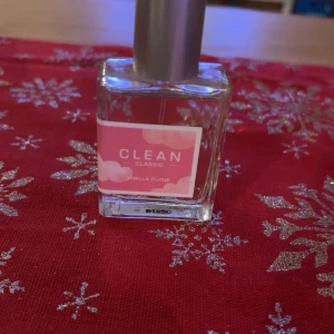 Clean Classic Vanilla Cloud Parfym - En elegant parfymflaska från Clean med doften Vanilla Cloud. Flaskan är fyrkantig och genomskinlig med en silverfärgad kork. Etiketten är rosa med vita moln och texten 'Clean Classic'. Perfekt för den som älskar en mjuk och söt vaniljdoft.
