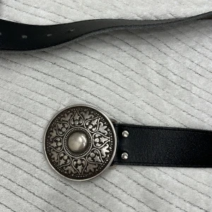 Svart bälte med dekorativt spänne - Snyggt svart bälte i skinn med ett stort, runt och detaljerat silverfärgat spänne. Perfekt för att ge en stilren touch till din outfit. Justerbar längd med flera hål.