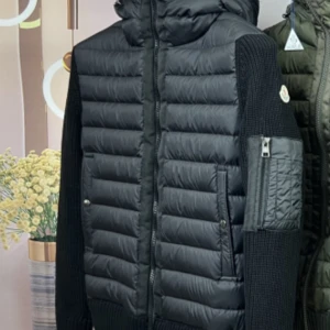 Svart pufferjacka från Moncler - Snygg svart pufferjacka från Moncler med quiltade paneler och stickade ärmar. Jackan har en huva och dragkedja framtill samt en praktisk ficka på ärmen. Perfekt för kyliga dagar!