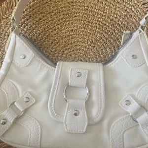 Vit axelväska med detaljer - Snygg vit axelväska med dekorativa spännen och dragkedja. Väskan har en stilren design med krokodilmönstrade detaljer och metallspännen. Perfekt för att ge din outfit en chic touch.