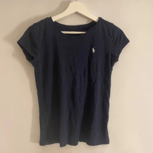 Mörkblå t-shirt från Ralph Lauren - Snygg mörkblå t-shirt från Ralph Lauren med korta ärmar och klassisk passform. Den har en liten broderad logotyp på bröstet i vitt. Perfekt för en stilren och avslappnad look.