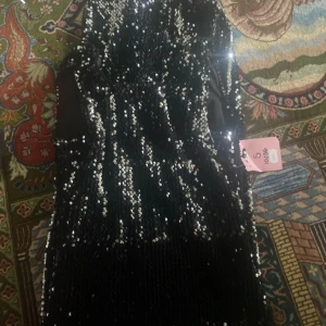 Svart paljettklänning - Snygg svart klänning täckt med glittriga paljetter. Perfekt för en festlig kväll med sin eleganta design och glansiga yta. Klänningen har en figurnära passform som framhäver silhuetten.